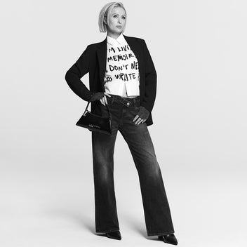 Person wearing a black blazer, white shirt with text, and dark jeans on a light gray background