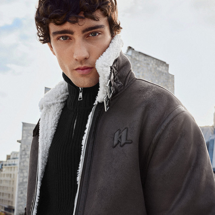 Man wearing a brown jacket with white fur lining, standing against an urban skyline.